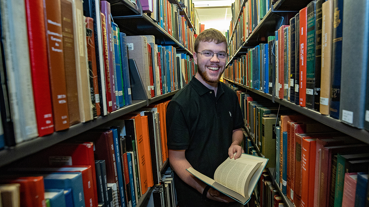SIU linguistics student Noah Clark stands between two bookshelves in the library holding book